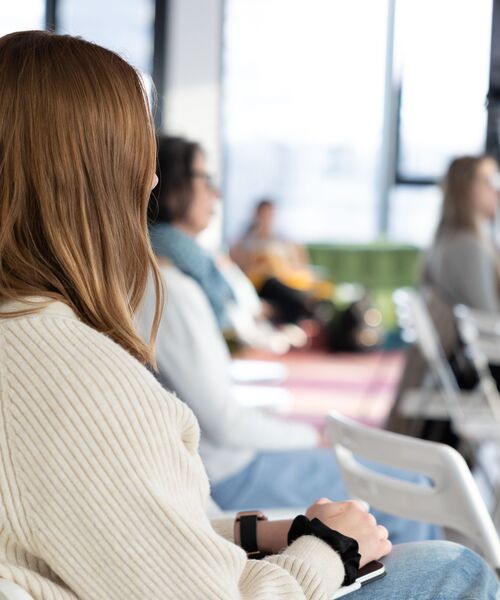 Person sitzt in heller Umgebung auf einem Stuhl und hört aufmerksam zu, im Hintergrund weitere Teilnehmende in unscharfer Ansicht.