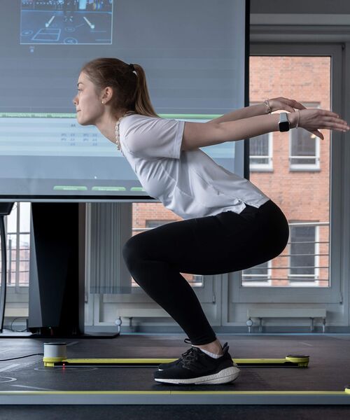 Junge Frau führt eine sportliche Übung im Labor mit Bewegungsanalyse durch, im Hintergrund läuft eine digitale Auswertung.
