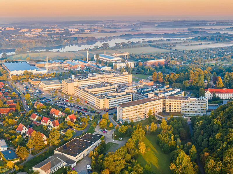 Luftaufnahme eines weitläufigen Gebäudekomplexes inmitten grüner Landschaft, umgeben von Wohnhäusern, Straßen und Feldern.