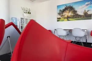 Moderner Warteraum mit roten Designerstühlen, weißen Möbeln und einem großen Wandbild eines Baumes in der Sonne.