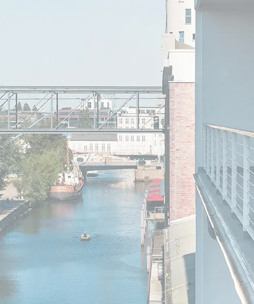 Brücke aus Stahl und Glas verbindet zwei Gebäude über einem Kanal, im Hintergrund Wasser und Stadtansicht.