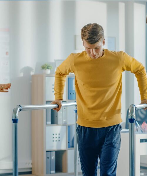 Ein junger Mann in gelbem Pullover macht Gehübungen an parallel verlaufenden Handläufen in einem Rehabilitationsraum.