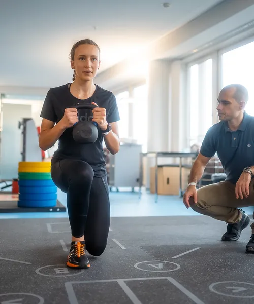 Person trainiert mit einer Kettlebell unter Anleitung eines Trainers in einem modernen Fitness- oder Rehabilitationsraum.