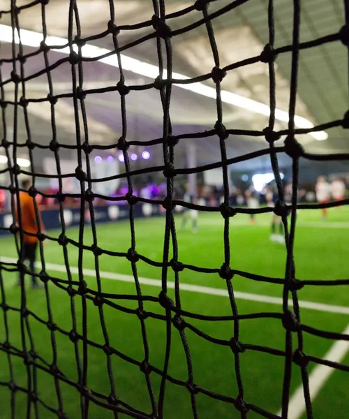 Blick durch ein Fußballnetz auf ein Hallenspielfeld mit Spielern und beleuchteter Kulisse im Hintergrund.