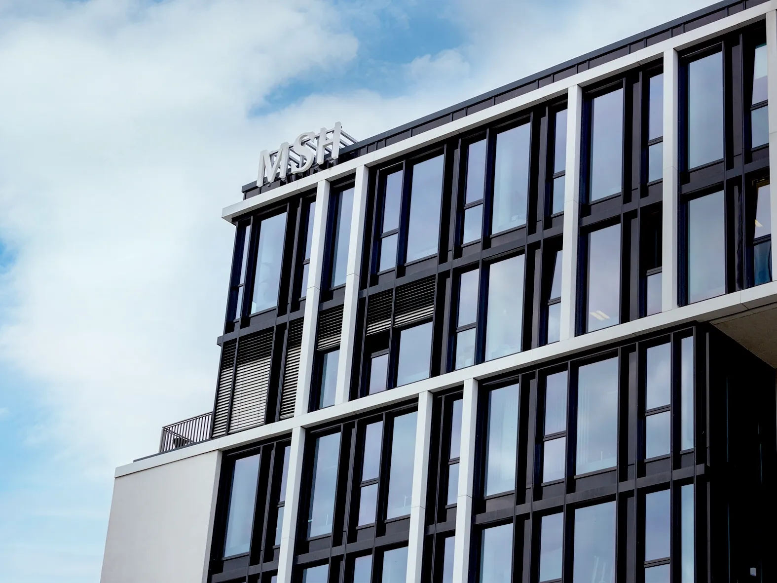 Fassade eines modernen Bürogebäudes mit großen Glasfenstern und klaren Linien vor teils bewölktem Himmel.