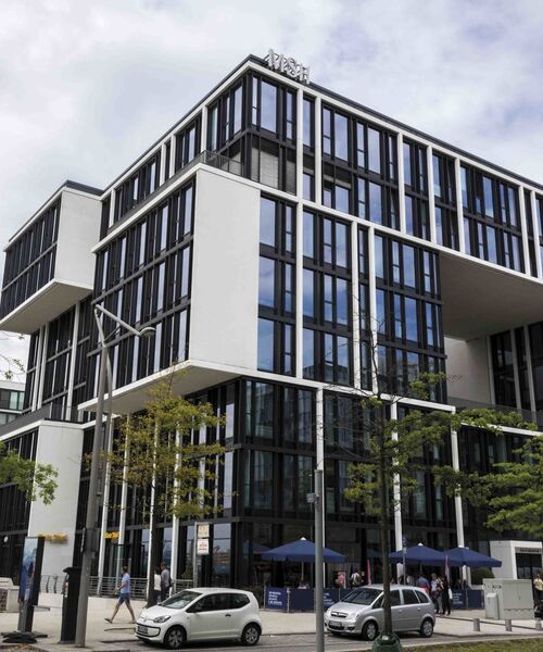 Modernes Bürogebäude mit markanter Glasfassade und auskragenden Bauelementen, davor Bäume, Autos und Passanten auf der Straße.