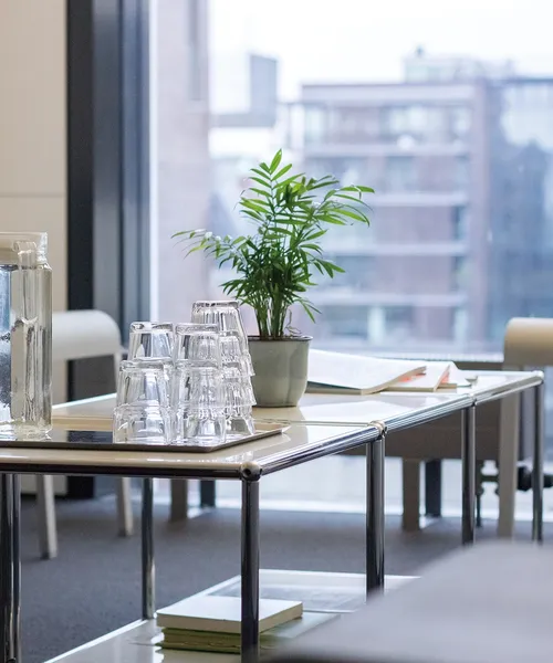 Glaskaraffe und gestapelte Trinkgläser auf einem Tisch neben einer kleinen Pflanze und geöffneten Heften in einem hellen Raum mit Stühlen.