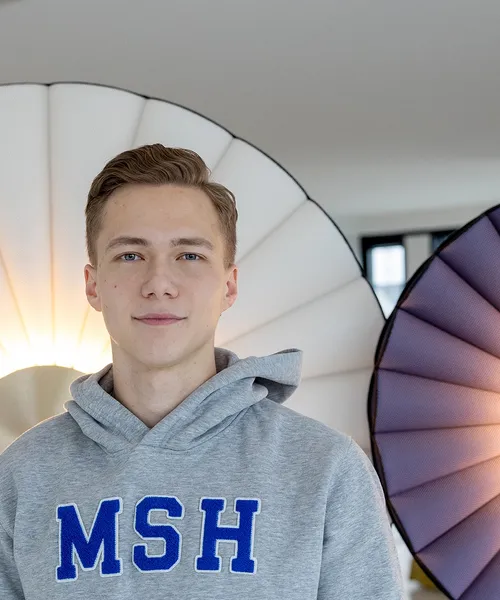 Ein junger Mann im grauen Hoodie steht zwischen zwei großen, fächerförmigen Leuchten, die im Hintergrund warm leuchten.