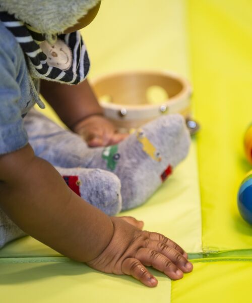 Kleinkind sitzt auf einer gelben Spielmatte, greift nach einem Spielzeug und hat eine kleine Trommel neben sich liegen.