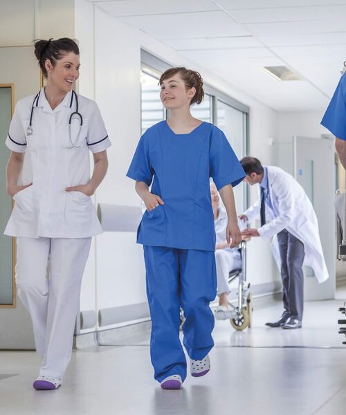 Zwei medizinische Fachkräfte in blauer und weißer Kleidung gehen lächelnd einen Krankenhausflur entlang, während im Hintergrund ein Arzt einem Patienten im Rollstuhl hilft.