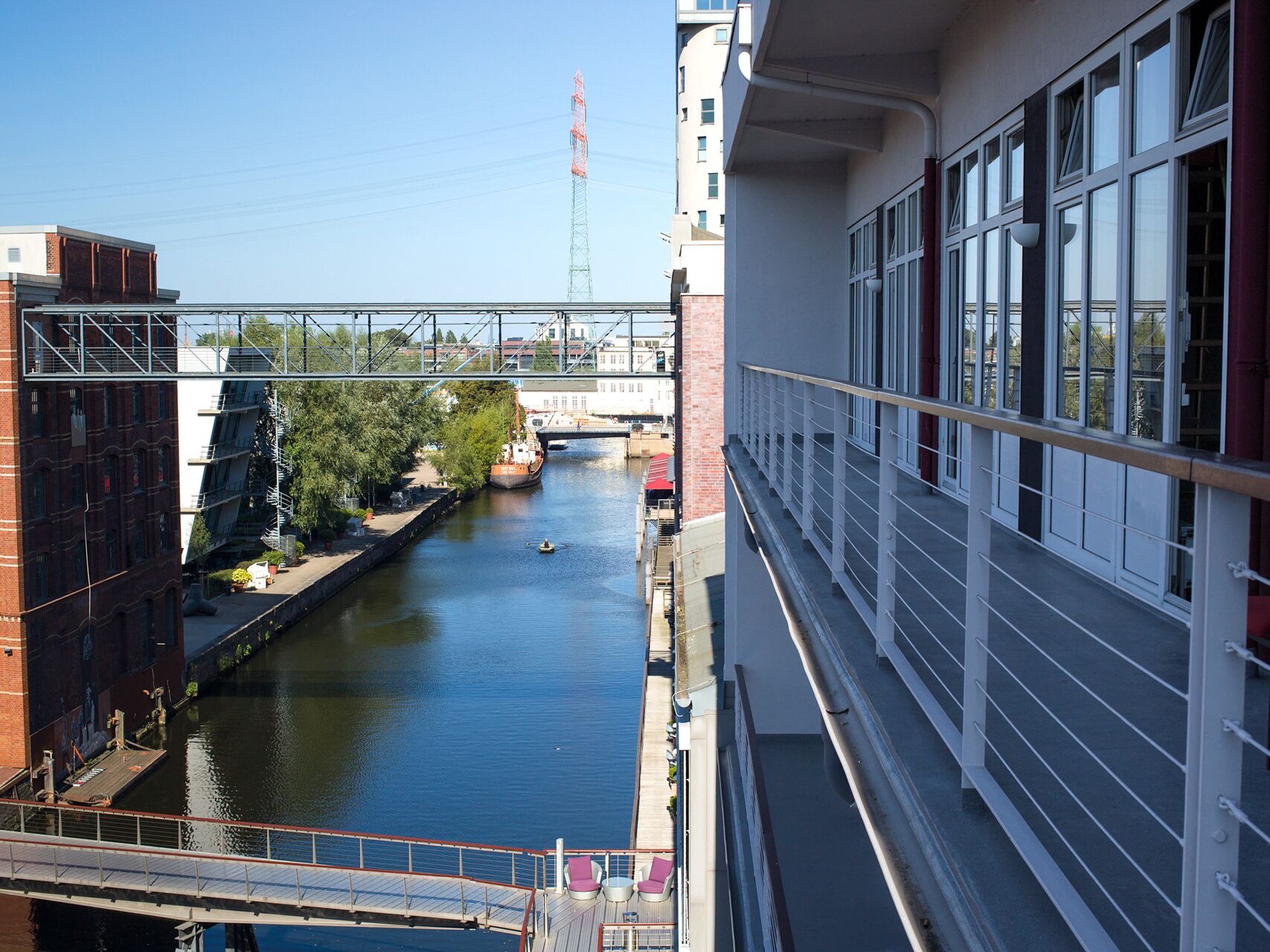 Blick von einem Gebäude auf einen Kanal mit klar blauem Wasser, flankiert von modernen Häusern und einer Brücke im Hintergrund. Ein Boot und ein Kajak sind auf dem Wasser zu sehen.