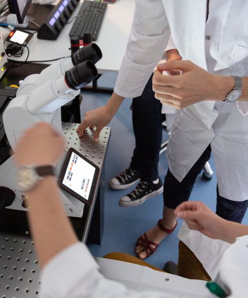 Personen in Laborkitteln arbeiten gemeinsam an einem Mikroskop mit digitaler Anzeige.