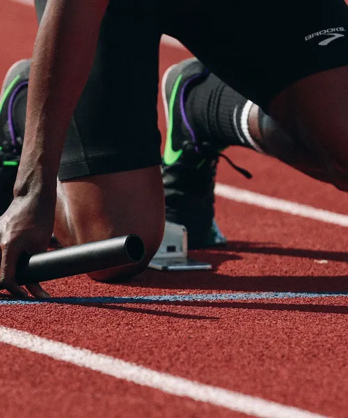 Nahaufnahme eines Sprinters in Startposition auf der Laufbahn mit Staffelstab in der Hand.