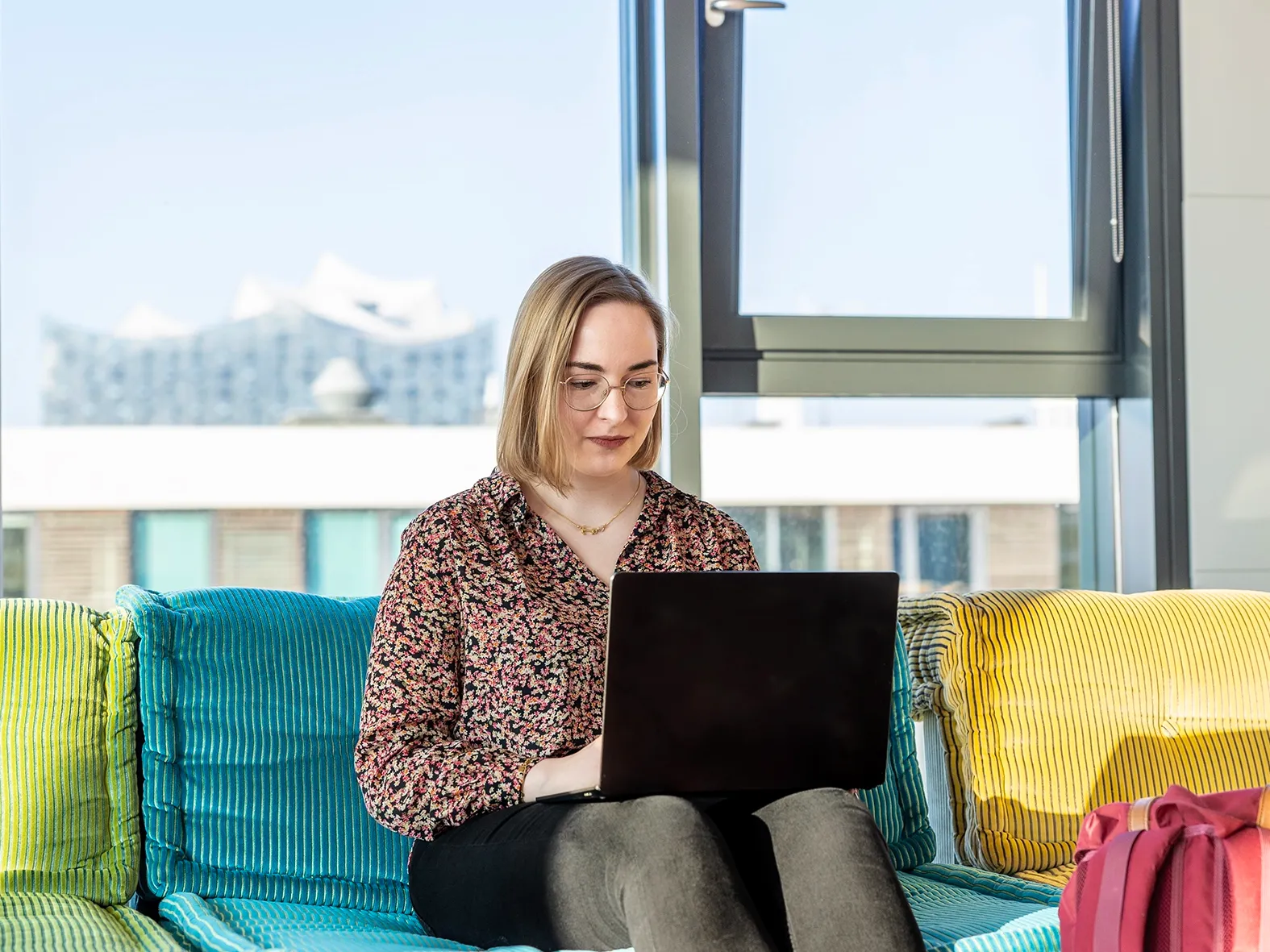Frau mit Brille sitzt auf einem Sofa und arbeitet konzentriert an einem Laptop.