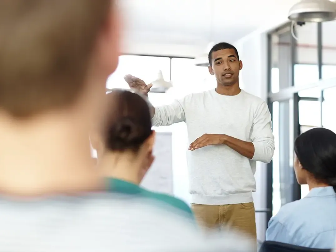 Ein junger Mann hält einen Vortrag vor einer kleinen Gruppe und gestikuliert mit den Händen, um etwas zu erklären.
