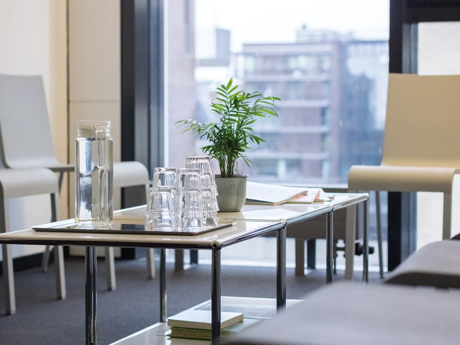 Glaskaraffe und gestapelte Trinkgläser auf einem Tisch neben einer kleinen Pflanze und geöffneten Heften in einem hellen Raum mit Stühlen.