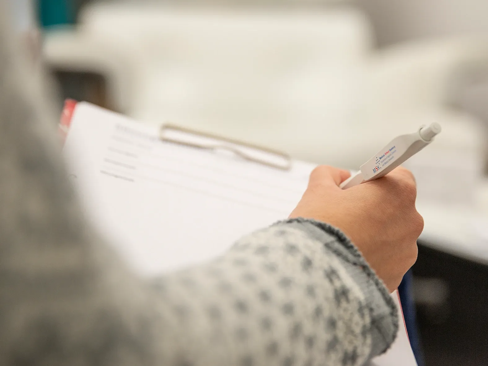 Hand hält einen Stift über einem Klemmbrett mit Papier, bereit zum Schreiben oder Ausfüllen eines Formulars.