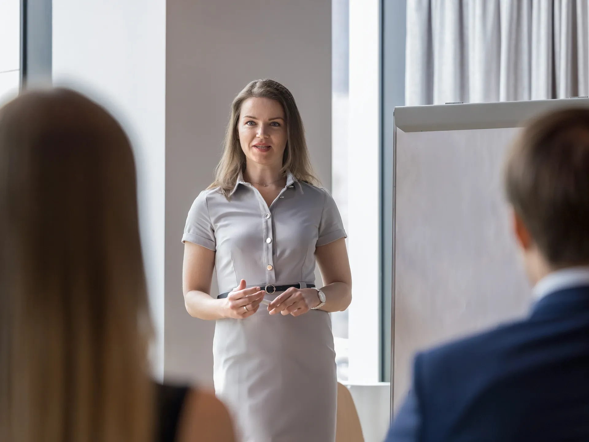 Eine Frau in grauem Kleid spricht vor zwei zuhörenden Personen in einem Besprechungsraum.