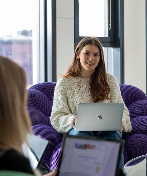 Junge Frau sitzt mit einem Laptop auf einem lila Sessel und lächelt, im Vordergrund eine weitere Person mit Laptop.