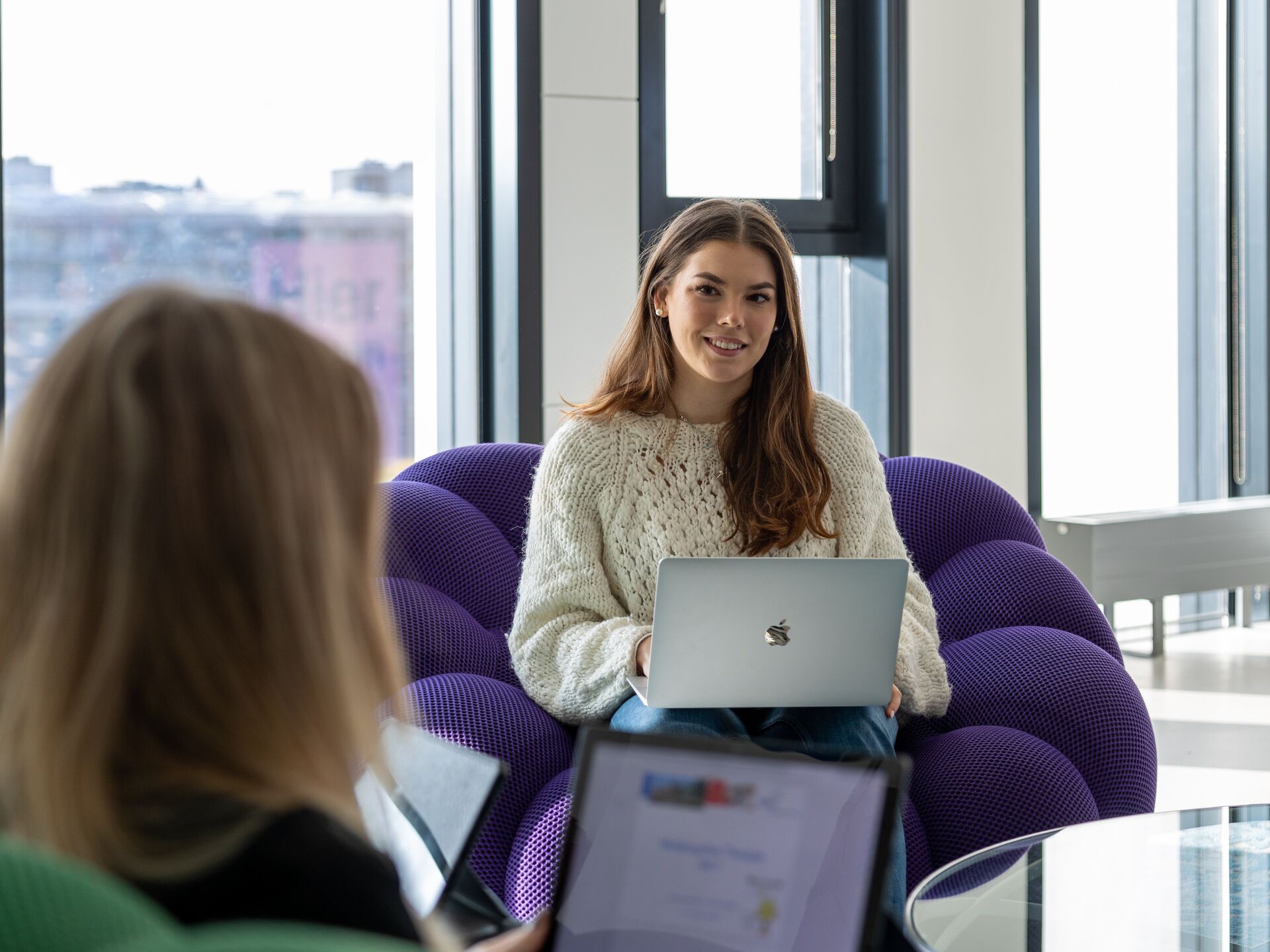 Junge Frau sitzt mit einem Laptop auf einem lila Sessel und lächelt, im Vordergrund eine weitere Person mit Laptop.