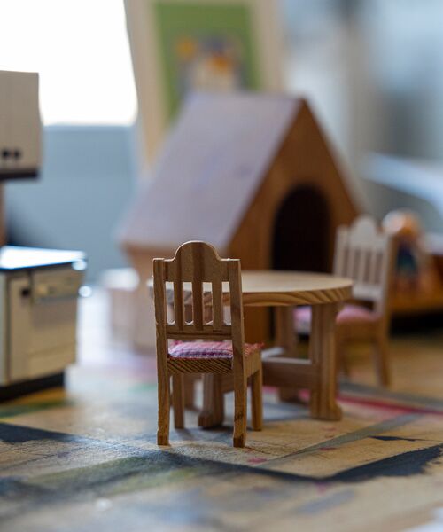 Miniaturmöbel mit einem kleinen Holztisch und zwei Stühlen stehen auf einem bunt gemusterten Untergrund, im Hintergrund ist ein Puppenhaus zu sehen.