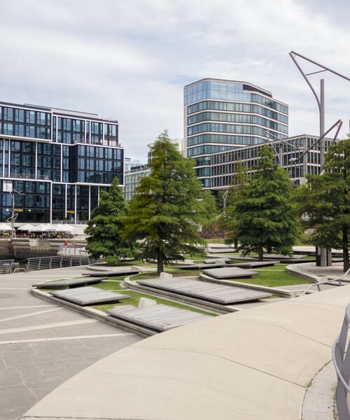Moderne Uferpromenade mit Sitzbänken, Grünflächen und Bäumen, im Hintergrund moderne Bürogebäude aus Glas und Stahl.