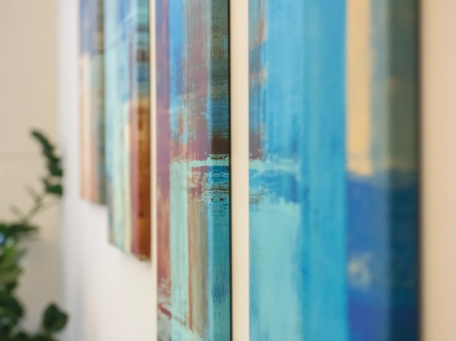 Nahaufnahme farbenfroher abstrakter Gemälde an einer Wand, im Hintergrund eine grüne Pflanze in heller Umgebung.