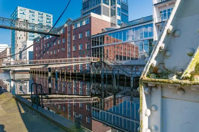 Moderne Gebäudefront am Wasser mit Backstein- und Glasfassaden, deren Spiegelung sich klar in der ruhigen Wasseroberfläche zeigt.