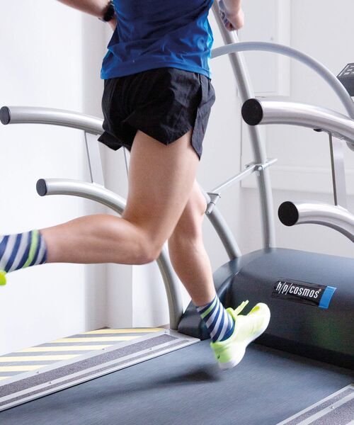 Person läuft in Sportsachen auf einem Laufband, Fokus auf den Beinen und Laufschuhen.