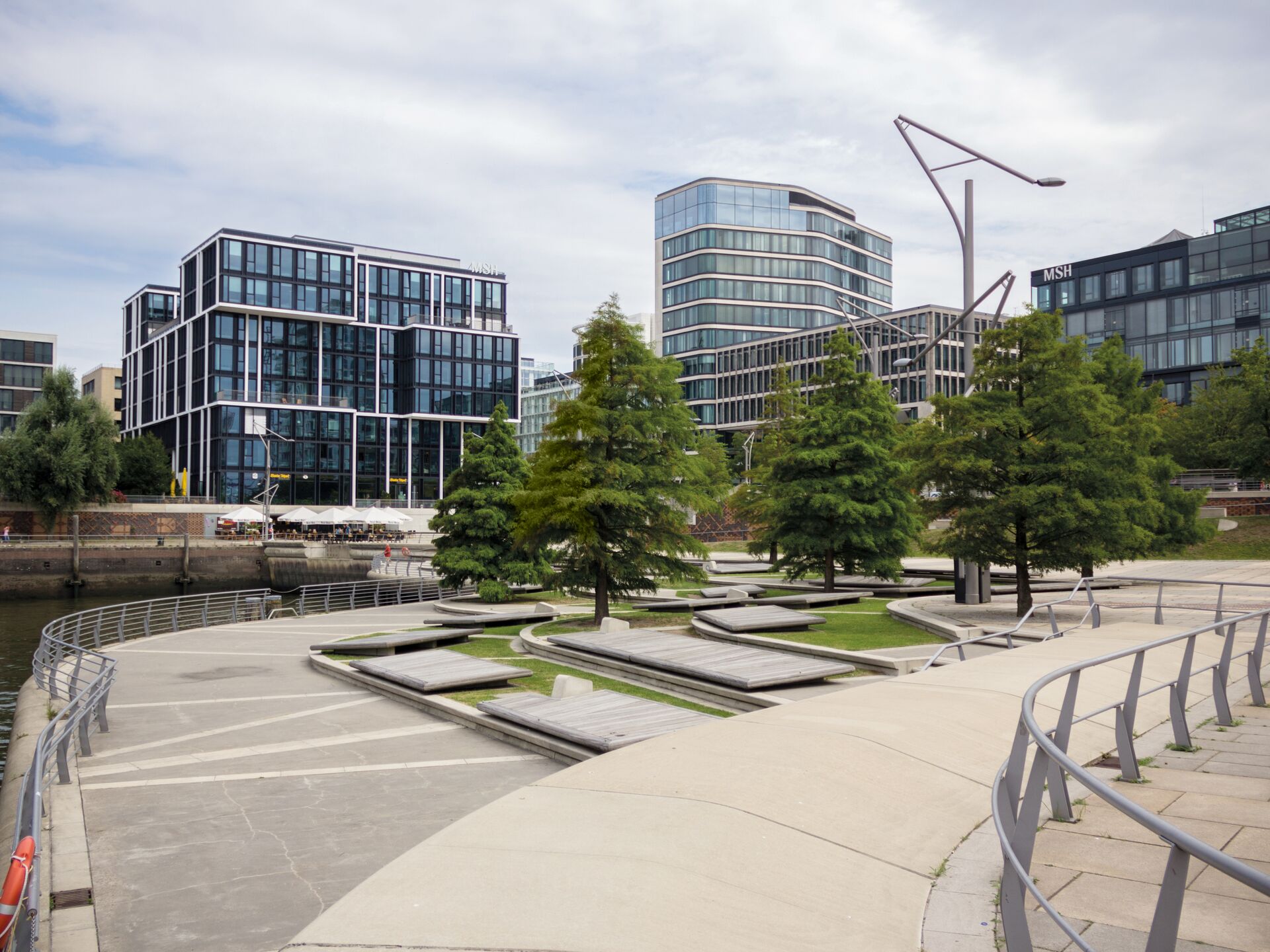 Moderne Uferpromenade mit Sitzbänken, Grünflächen und Bäumen, im Hintergrund moderne Bürogebäude aus Glas und Stahl.