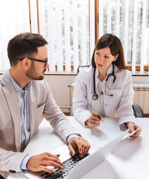 Ein Arztgespräch: Eine Ärztin mit Stethoskop erklärt einem Mann im Anzug etwas, während er an einem Laptop arbeitet.