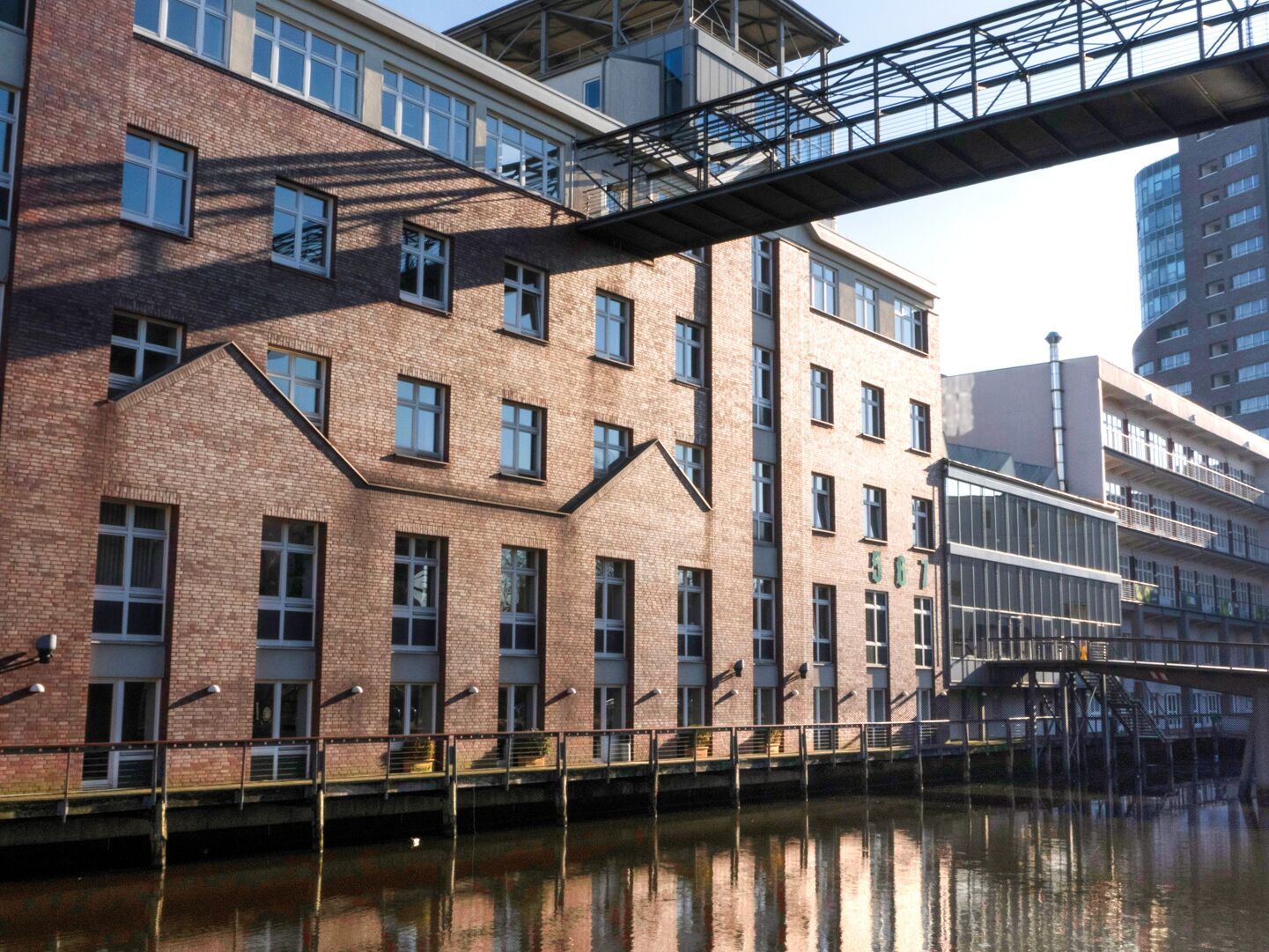 Ziegelrotes Gebäude mit mehreren Stockwerken und großen Fenstern, das direkt an einem Wasserlauf liegt; darüber verläuft eine verglaste Fußgängerbrücke, die zwei Gebäudeteile verbindet.