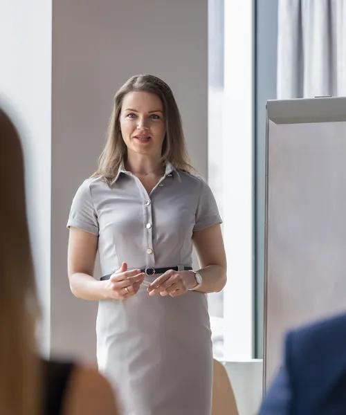 Eine Frau in grauem Kleid spricht vor zwei zuhörenden Personen in einem Besprechungsraum.