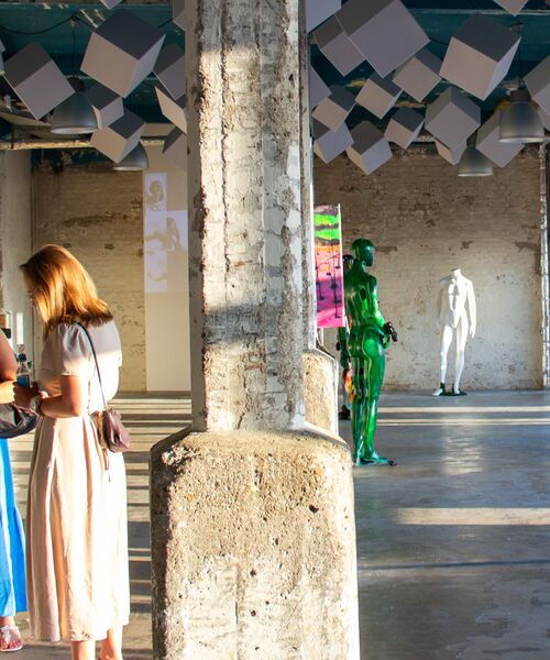 Menschen in einer hellen Kunstausstellung mit Betonpfeilern, farbigen Skulpturen und moderner Rauminstallation unter der Decke.