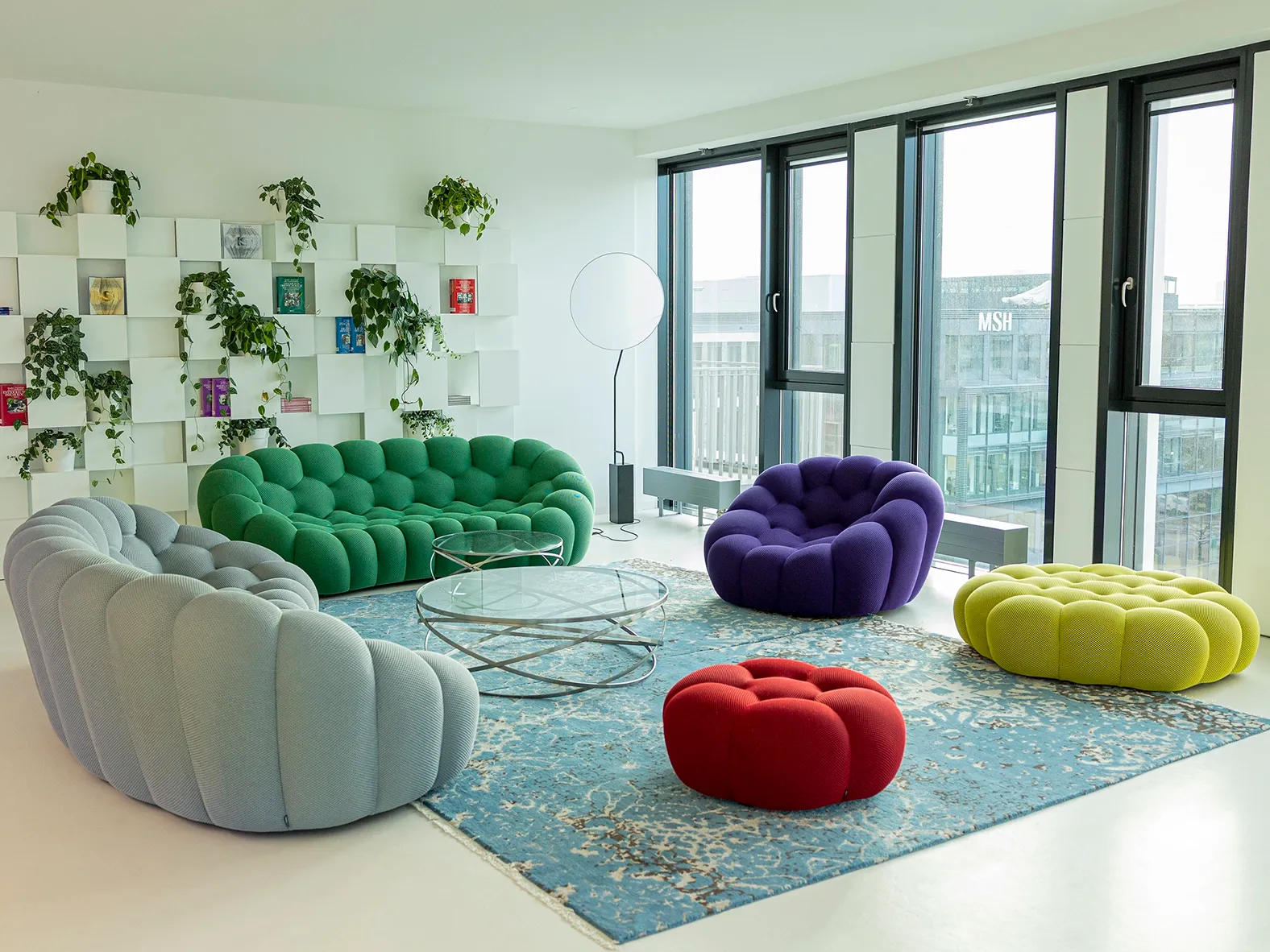 Moderner Aufenthaltsraum mit bunten Designersofas, Glas­tischen und großen Fenstern, durch die Tageslicht einfällt – eine gemütliche und offene Atmosphäre.