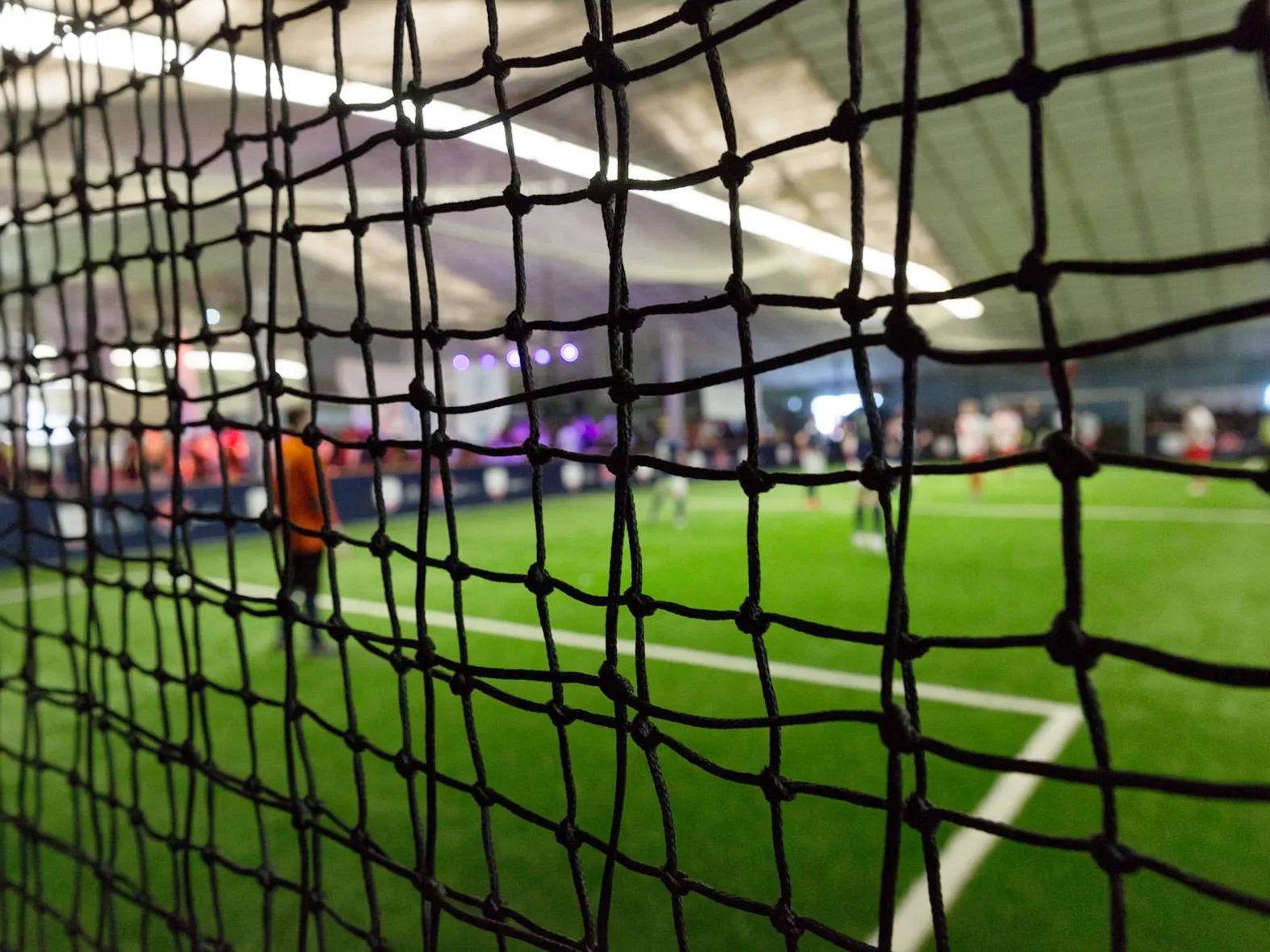 Blick durch ein Fußballnetz auf ein Hallenspielfeld mit Spielern und beleuchteter Kulisse im Hintergrund.