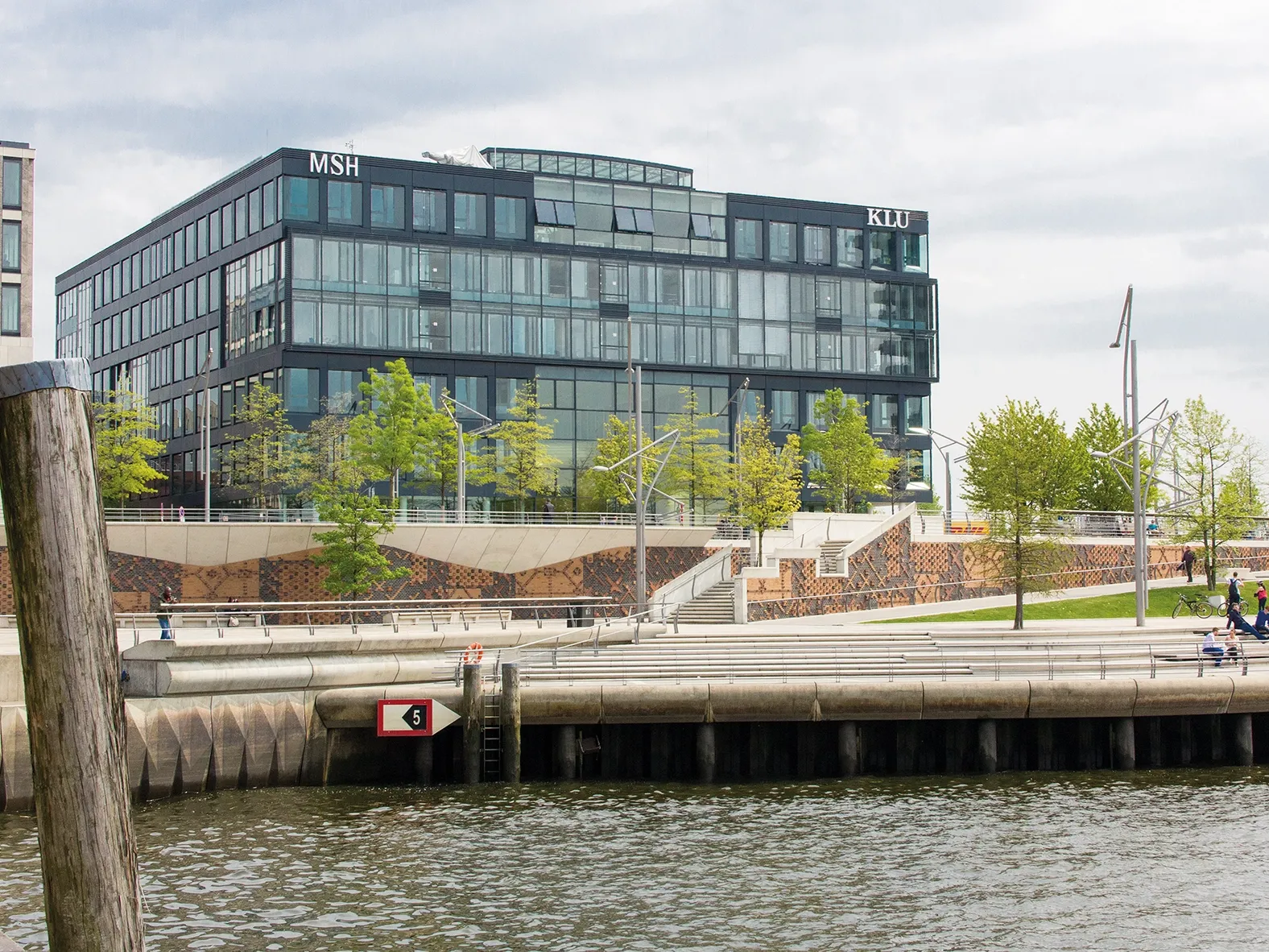 Modernes Bürogebäude mit Glasfassade am Wasser, umgeben von Bäumen, Promenade und Spaziergängern.