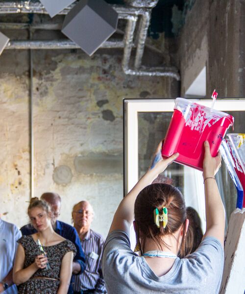 Person gießt leuchtend pinke Farbe auf eine Leinwand, während mehrere Menschen im Hintergrund aufmerksam zusehen.
