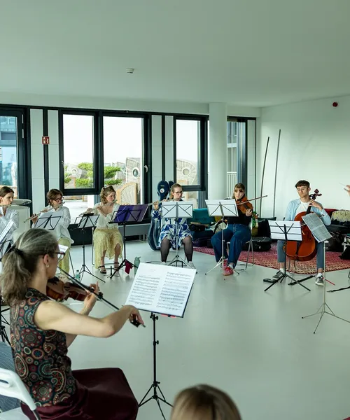 Musikensemble mit verschiedenen Instrumenten probt in einem hellen Raum, geleitet von einer Dirigentin in grüner Kleidung.