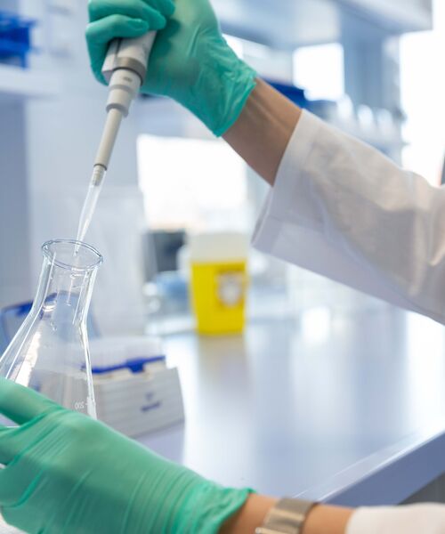 Person im Labor befüllt mit einer Pipette einen Erlenmeyerkolben, trägt Handschuhe und Laborkittel.
