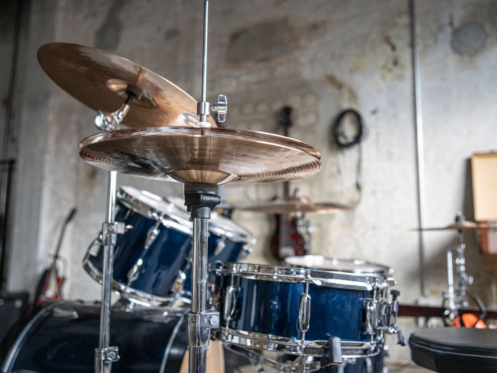 Ein Schlagzeug mit blauen Trommeln und glänzenden Becken steht in einem Proberaum mit heller, leicht abgenutzter Wand.