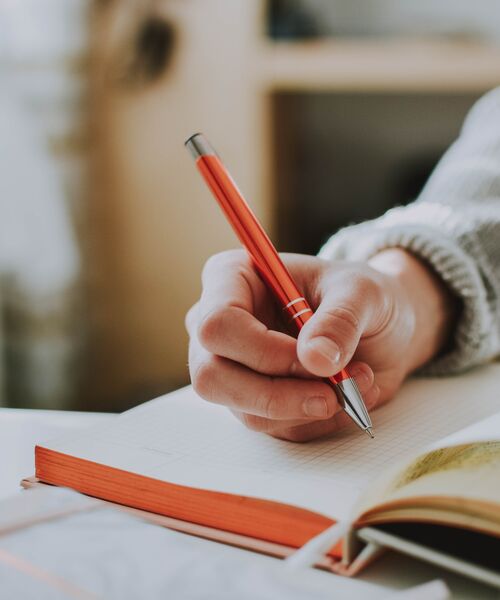 Hand hält einen roten Stift und schreibt in ein aufgeschlagenes Notizbuch.