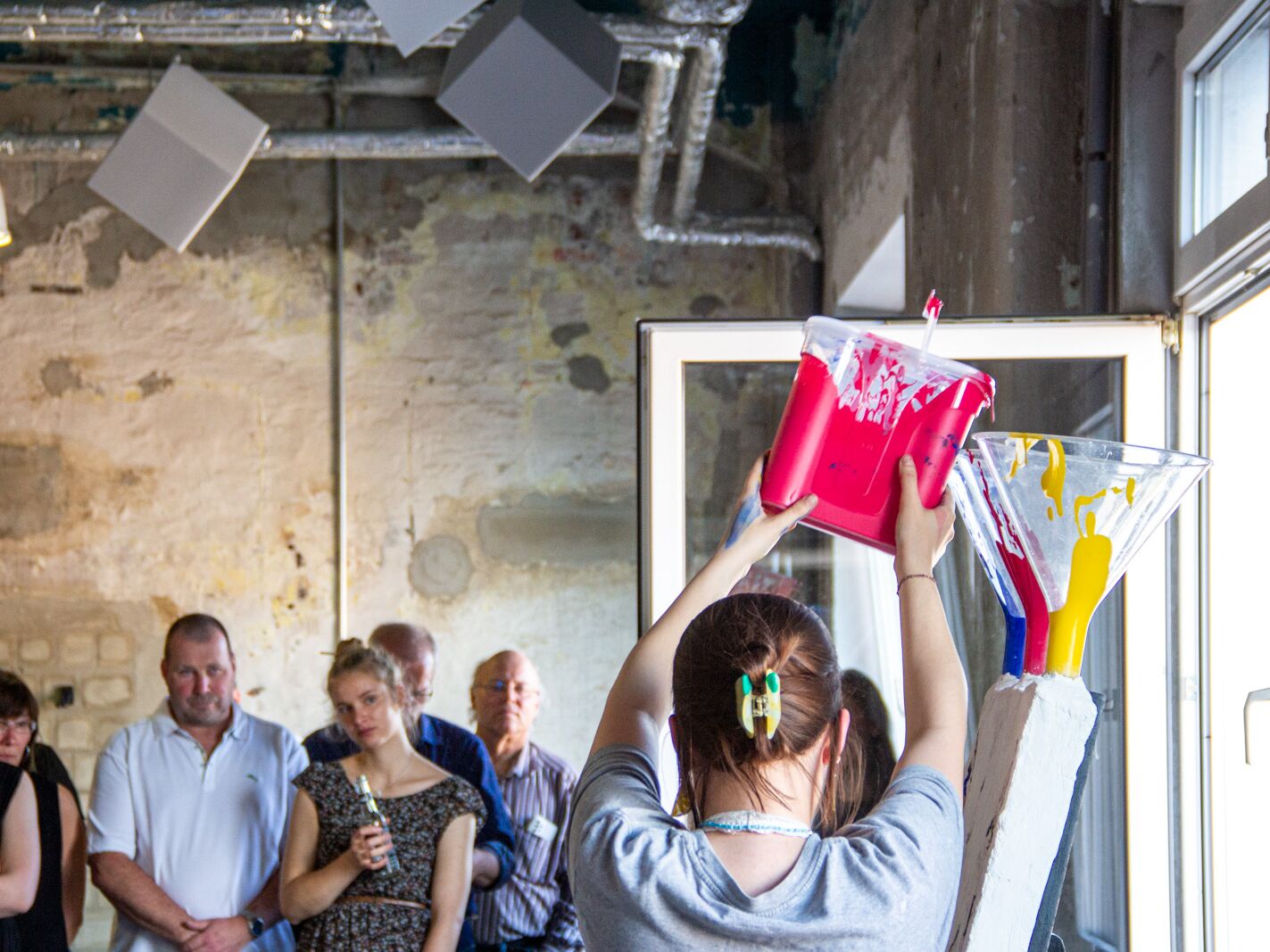Person gießt leuchtend pinke Farbe auf eine Leinwand, während mehrere Menschen im Hintergrund aufmerksam zusehen.