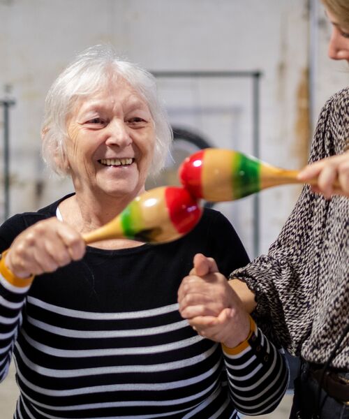 Ältere Frau lächelt und schüttelt bunte Maracas, während sie von einer anderen Person unterstützt wird.