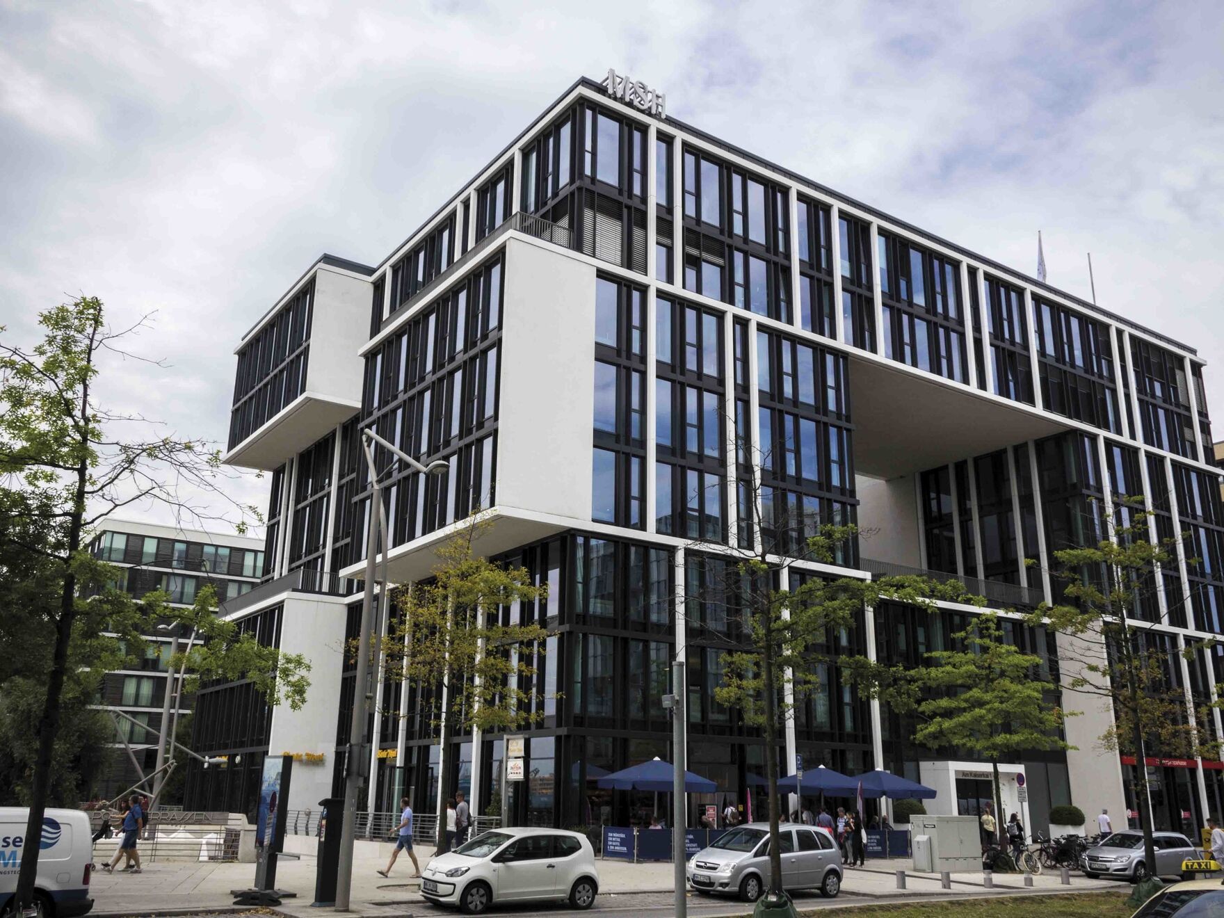 Modernes Bürogebäude mit markanter Glasfassade und auskragenden Bauelementen, davor Bäume, Autos und Passanten auf der Straße.