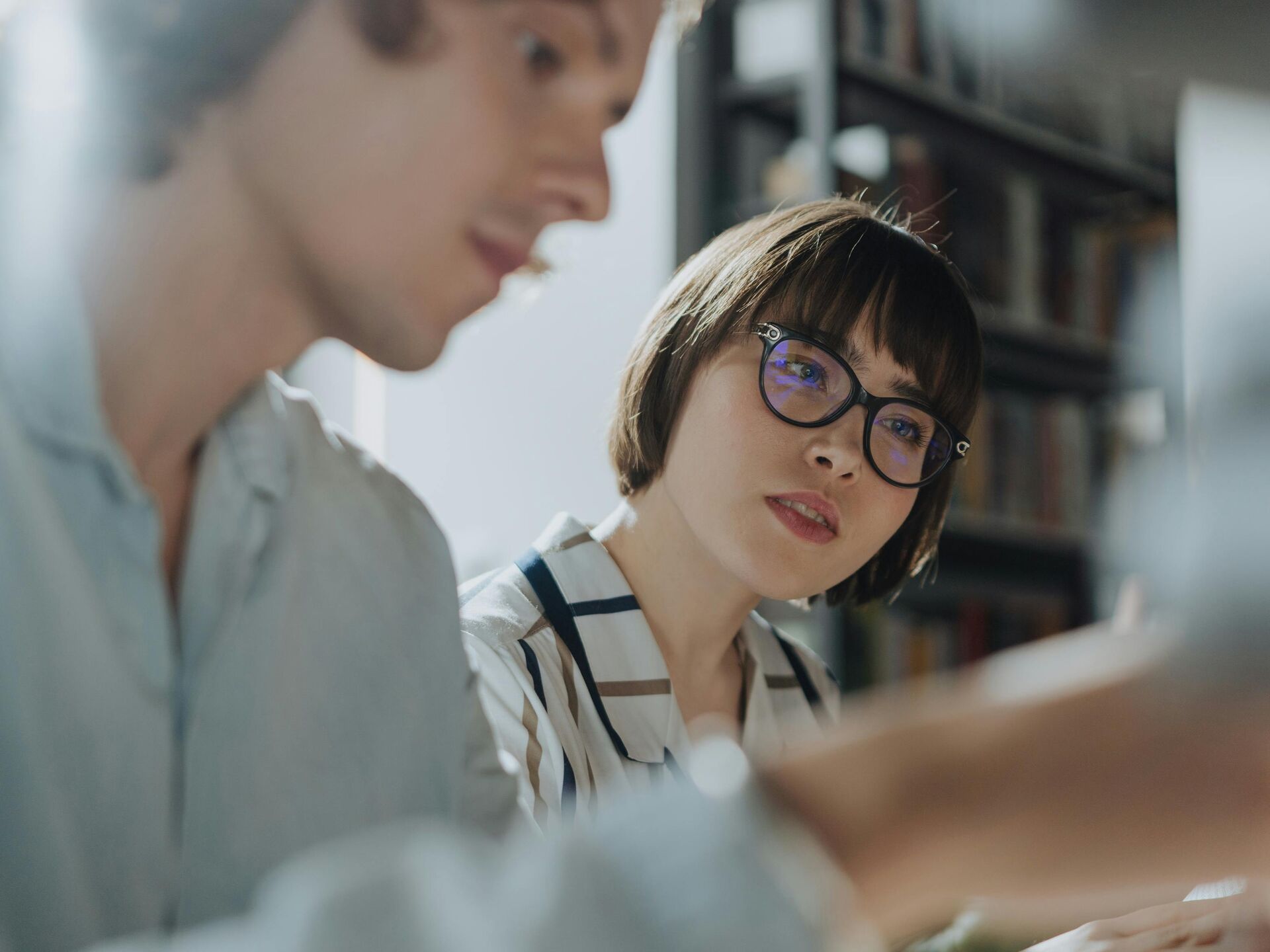 Person mit Brille hört aufmerksam einem Gesprächspartner zu, im Hintergrund sind Bücherregale zu sehen.