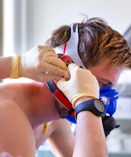 Person mit Atemmaske wird von einer Fachkraft mit Handschuhen für eine sportmedizinische Untersuchung vorbereitet.