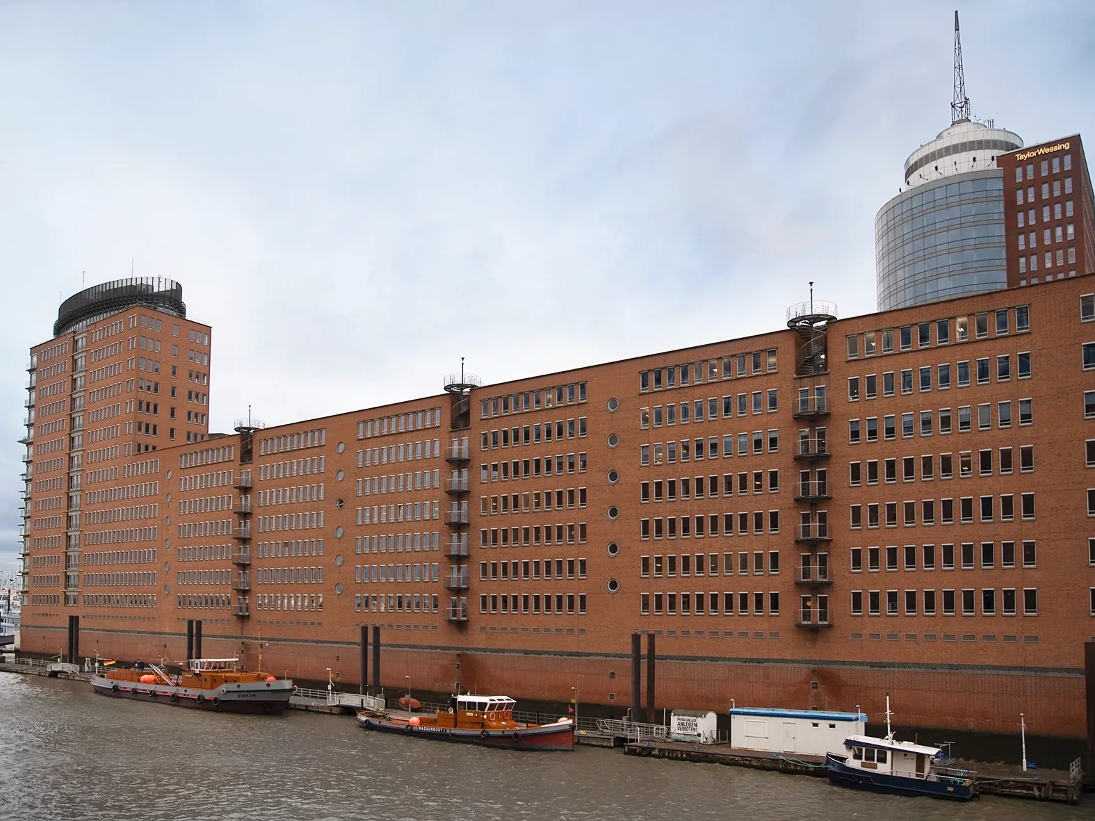 Großes Backsteingebäude mit zahlreichen Fenstern direkt am Wasser, davor mehrere Boote am Ufer festgemacht.
