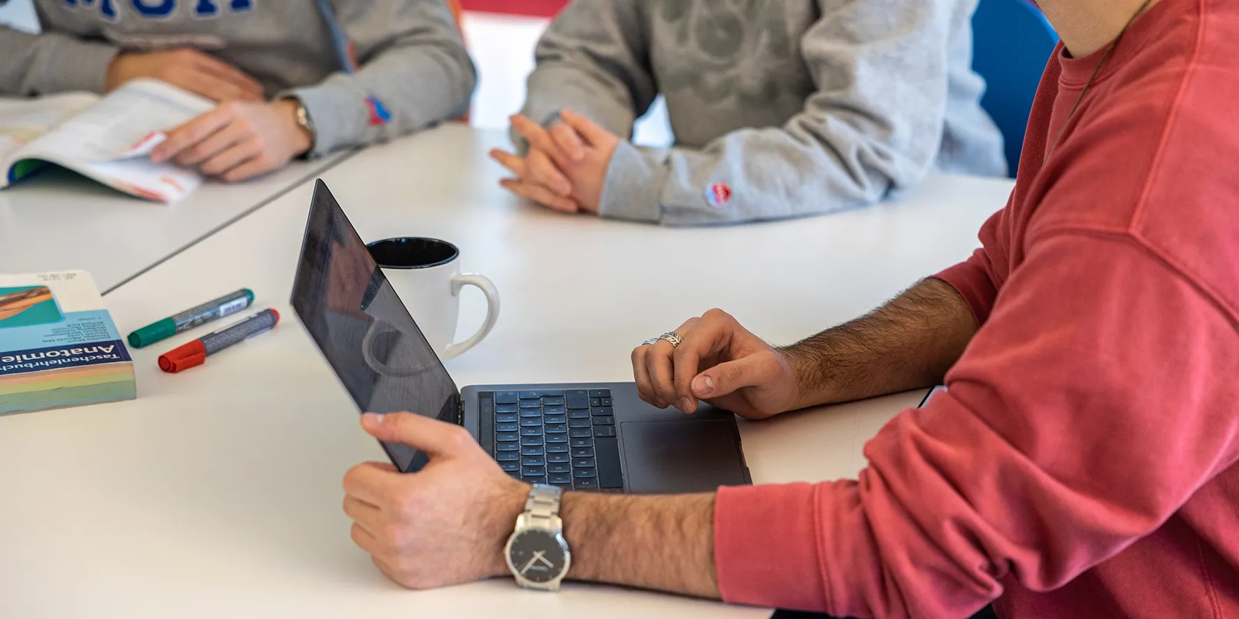 Studierende arbeiten gemeinsam an einem Tisch mit Laptop, Tasse, Stiften und einem Anatomiebuch.