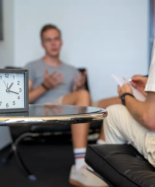 Nahaufnahme einer Uhr auf einem Tisch, im Hintergrund Gespräch zwischen zwei Personen in einer Beratungssituation.